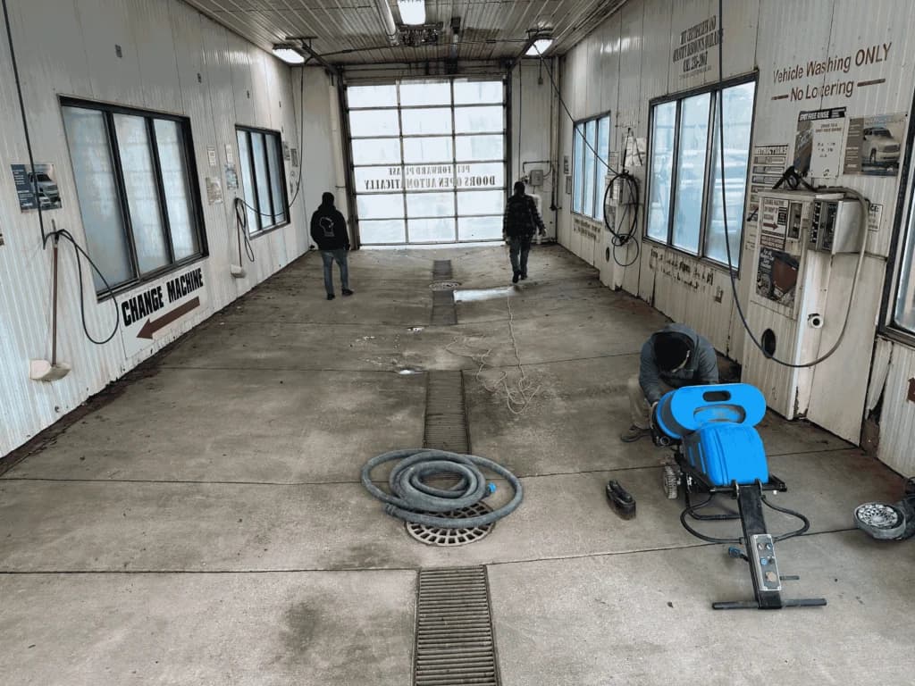 Technician using a blue floor scrubber to clean and prep a concrete floor inside a vehicle wash/service bay.