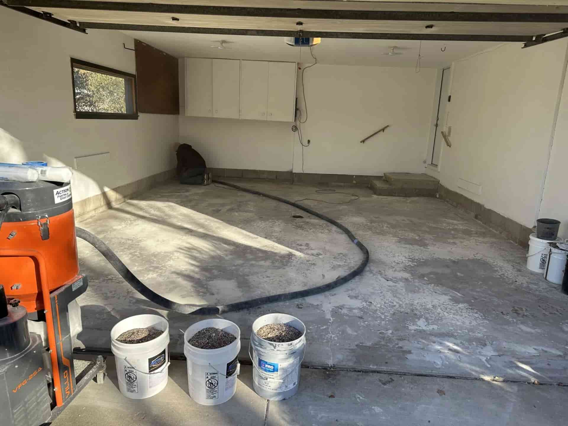 Garage interior with concrete floor preparation underway using floor grinder, dust-control vacuum, and coating materials staged for application.